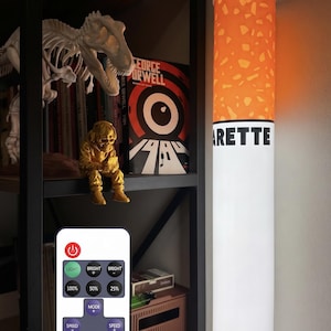 May include: A cylindrical floor lamp designed to resemble a cigarette, featuring an orange filter section with a speckled pattern and a white body with the black text "ARETTE" visible. The lamp is accompanied by a white remote control with buttons.