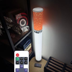 May include: A cylindrical floor lamp designed to resemble a cigarette, featuring a white base and an orange filter-patterned top with the partial text "GARETTE" printed in black. The lamp is accompanied by a small rectangular remote control.