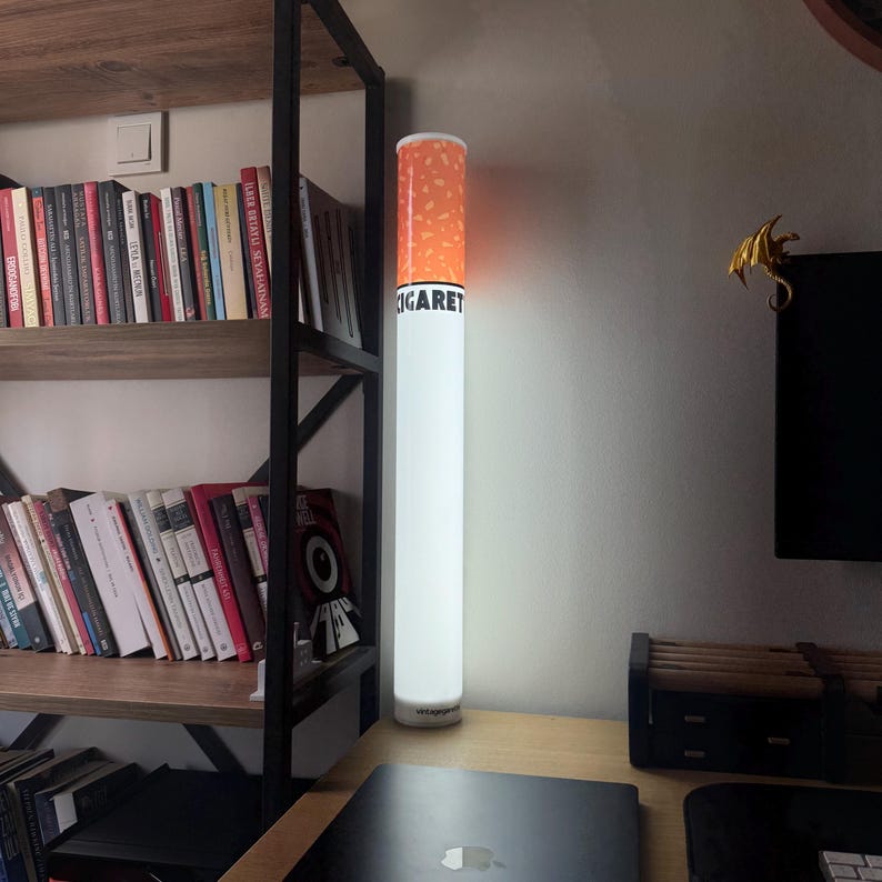May include: A novelty floor lamp shaped like a cigarette. The lamp has a white body with an orange and brown tip, and the word "CIGARET" is printed on the side. It stands tall on a desk near a bookshelf.