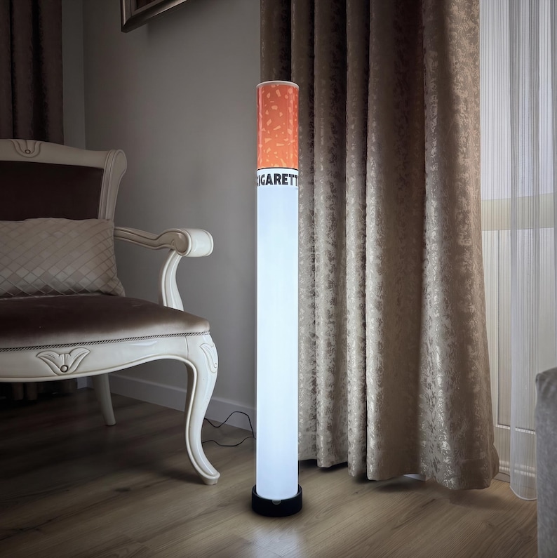 May include: A novelty floor lamp designed to resemble a cigarette. The lamp has a white, cylindrical body with a black base. The top section is orange, mimicking the lit end of a cigarette. The word "CIGARETT" is printed on the side.