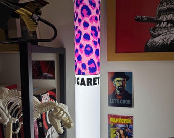 May include: A tall, cylindrical floor lamp shaped like a cigarette, featuring a pink and blue leopard print pattern at the top and the partial black text "IGARET" printed on a white background below the filter section, emitting a soft white glow.