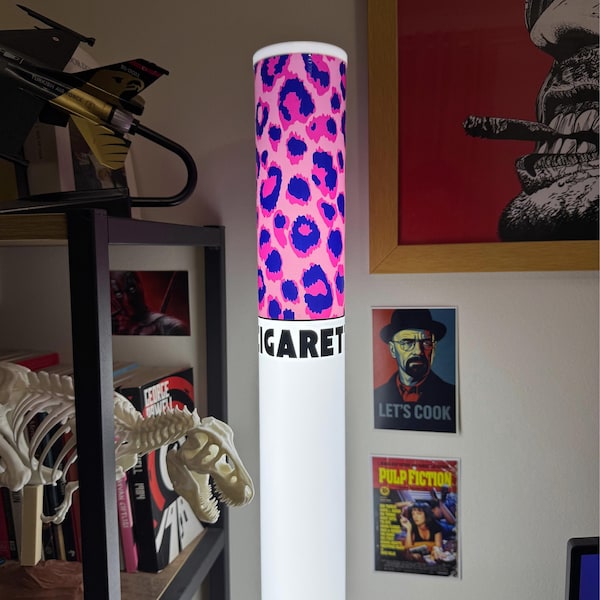 May include: A tall, cylindrical floor lamp shaped like a cigarette, featuring a pink and blue leopard print pattern at the top and the partial black text "IGARET" printed on a white background below the filter section, emitting a soft white glow.