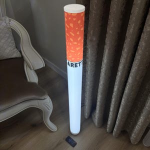 May include: A tall, cylindrical floor lamp shaped like a cigarette, featuring an orange filter section with a speckled pattern and a white body section. The word "GARETTE" is printed in black letters on the white portion. A small remote control lies on the floor.