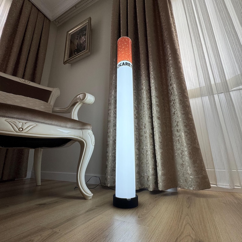 May include: A tall, novelty floor lamp shaped like a cigarette. The lamp has a white, cylindrical body with an orange and brown tip. The word "CIGARET" is printed on the side. The lamp stands on a black base and is placed on a wooden floor.