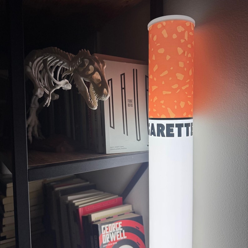 May include: A novelty floor lamp designed to resemble a giant cigarette. The lamp features a white cylindrical base with the word "CIGARETTE" printed on it, topped with an orange and white speckled design. A dinosaur skeleton and books are on a shelf in the background.