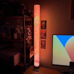 May include: A tall, cylindrical floor lamp with a warm, orange-red glow. The lamp features a printed design resembling a cigar wrapper with the text "VINTAGEGARETTO PREMIUM CIGARS ORIGINAL" and a central lion emblem, creating a textured appearance.