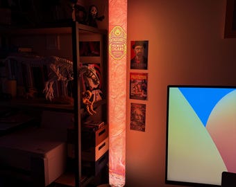 May include: A tall, cylindrical floor lamp emitting a warm, orange-red glow. The lamp features a printed design resembling a cigar wrapper with the text "VINTAGEGARETTO PREMIUM CIGARS ORIGINAL" and a central lion emblem, creating a textured appearance.