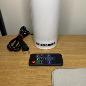 May include: A white cylindrical lamp with the text VINTAGEGARETTE printed in black near the base. The lamp includes a black power cord with a two-pin plug and a rectangular black remote control featuring buttons for power, brightness, and speed.