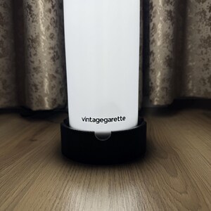 May include: A white cylindrical object with the text "vintagegarette" in black, resting on a black base. The background features a wooden surface and a patterned curtain.