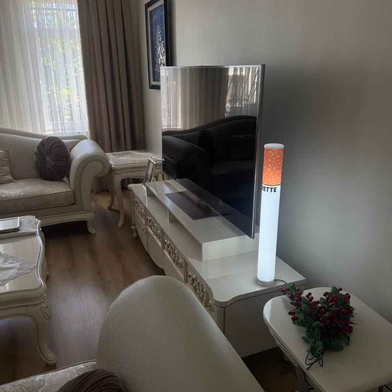 May include: A modern living room with a large flat-screen TV and a decorative lamp shaped like a cigarette. The lamp has an orange and white design with the word "ETTE" visible. The room also features white furniture and a small side table.