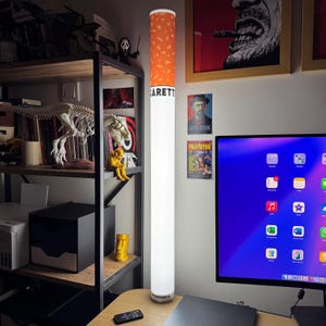 May include: A tall, cylindrical floor lamp designed to resemble a cigarette, featuring a white base and an orange, speckled filter top. The lamp displays the text "GARETTE" in black letters near the top and "VINTAGEGARETTE" at the bottom.