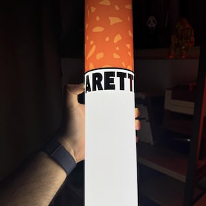 May include: A cylindrical object resembling a cigarette with a white base and an orange filter section featuring a speckled pattern. The word ARETT is printed in bold black capital letters across the white section near the orange filter area.