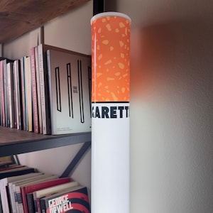 May include: A tall, cylindrical decorative object resembling a cigarette. The top portion is orange with a speckled pattern, transitioning to white with the word "GARETT" in black. The object is displayed near a bookshelf.
