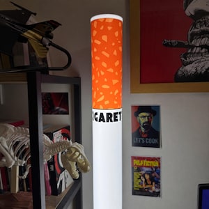 May include: A cylindrical floor lamp designed to resemble a cigarette, featuring a white base and an orange filter section with a speckled pattern. The word CIGARETTE is printed in bold black capital letters on the white section of the lamp.