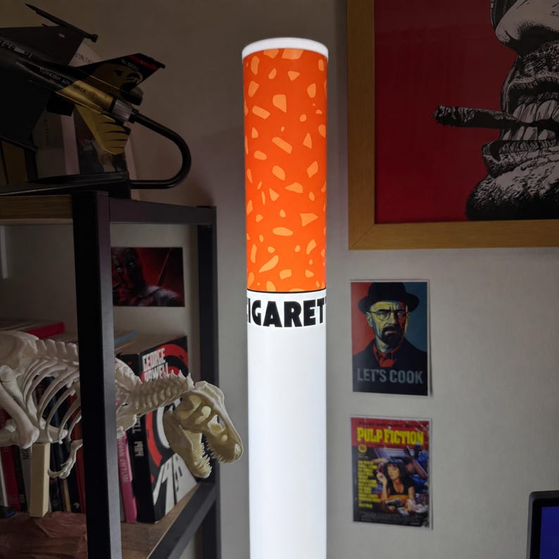 May include: A cylindrical floor lamp designed to resemble a cigarette, featuring a white base and an orange filter section with a speckled pattern. The word CIGARETTE is printed in bold black capital letters on the white section of the lamp.