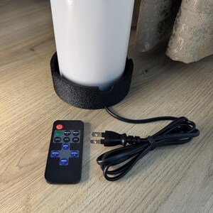 May include: A white cylindrical lamp with a black base, a remote control, and a power cord. The remote has buttons for power, brightness levels, and speed. The lamp is on a wooden surface.