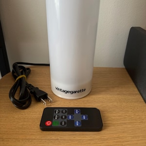 May include: A white cylindrical lamp with the text "vintagegarette" printed on the base. A black remote control with multiple buttons and a power cord are next to the lamp. The lamp is on a wooden surface.