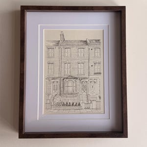 May include: A framed ink drawing on cream paper depicting a row of terraced houses with architectural details including bay windows, chimneys, and front doors. The artwork is enclosed in a dark brown wooden frame with a white mat border.