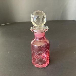 Può includere: Una piccola bottiglia in vetro rosa con un motivo a taglio di diamante sul corpo e un tappo in vetro trasparente, sferico e sfaccettato. La bottiglia presenta un collo corto e una base ampia e piatta, con una tonalità rosa traslucida.