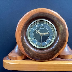 Puede incluir: Un reloj de sobremesa redondo de madera con un marco de vetas de madera marrón oscuro y bronceado claro. La esfera es negra con números romanos dorados y manecillas doradas ornamentadas. El texto "Jerger ANKER" está impreso en blanco.