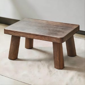 May include: A small, rectangular wooden stool with a dark brown finish and a visible natural wood grain texture. The stool features a flat top surface and is supported by four thick, sturdy, cylindrical legs that extend outward from the corners.