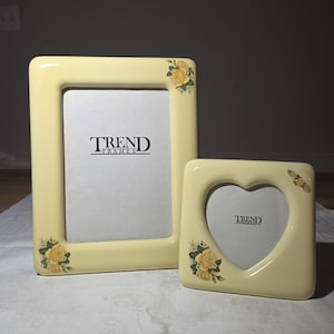 May include: Two cream-colored picture frames. One is rectangular, the other heart-shaped. Both have yellow rose accents in the corners. The rectangular frame has the text "TREND FRAMES" at the bottom. The heart-shaped frame also has the text "TREND FRAMES".