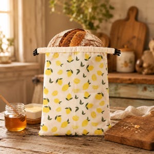 May include: A rectangular fabric bread bag with a cream background and a repeating pattern of yellow lemons and green leaves. The bag features a drawstring closure at the top and contains a round loaf of crusty, dark brown artisan bread.