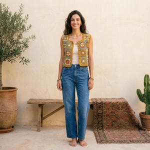 Può includere: Una donna indossa un gilet all'uncinetto giallo senape con motivi floreali quadrati nei toni del verde, arancione e bianco. Indossa jeans blu e un top bianco. Il gilet senza maniche ha uno scollo tondo.