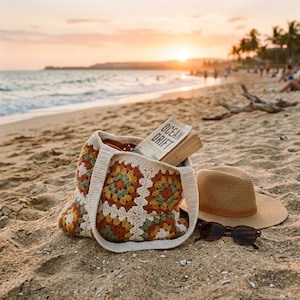 Pode incluir: Uma sacola tote de crochê com um padrão floral colorido, um chapéu de palha e óculos de sol repousam em uma praia arenosa. Um livro intitulado "Ocean Drift" é visível dentro da bolsa. A cena é banhada pela luz quente do pôr do sol.