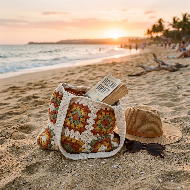 Puede incluir: Un bolso tote de crochet con un colorido estampado floral, un sombrero de paja y gafas de sol descansan en una playa de arena. Un libro titulado "Ocean Drift" es visible dentro de la bolsa. La escena est&aacute; ba&ntilde;ada por la c&aacute;lida luz de una puesta de sol.