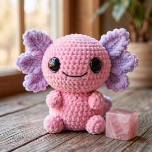 May include: A handmade pink and purple crochet axolotl plush toy with large black eyes and a smiling face. The plush is sitting on a wooden surface next to a pink rose quartz cube. The axolotl has detailed gills and a textured body.