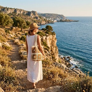 Puede incluir: Una persona con un vestido blanco, gafas de sol y un sombrero beige, lleva un bolso tote de ganchillo con cuadrados marrones y crema, caminando por un sendero costero. El fondo muestra el mar y los acantilados.