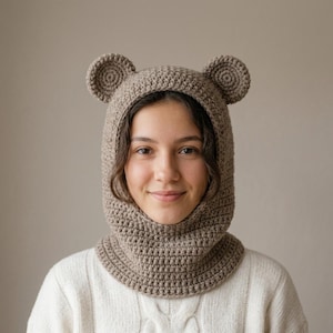 May include: A beige crochet balaclava featuring two rounded bear ears on top. The head covering is crafted from a textured yarn, designed to fit snugly around the head and neck, providing full coverage with a circular opening for the face.