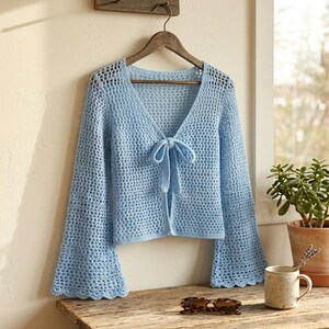 May include: A light blue, open-weave crochet cardigan with long, flared sleeves hangs on a wooden hanger. The cardigan features a V-neckline with a tie closure. The garment is displayed on a wooden surface with sunglasses and a mug.