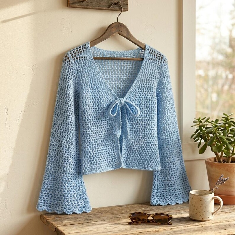 May include: A light blue, open-weave crochet cardigan with long, flared sleeves hangs on a wooden hanger. The cardigan features a V-neckline with a tie closure. The garment is displayed on a wooden surface with sunglasses and a mug.
