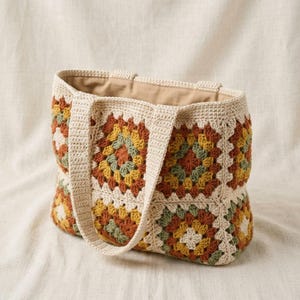 Puede incluir: Un bolso tote de crochet color crema con un colorido patr&oacute;n de cuadrados de la abuela en tonos naranja, amarillo y verde. El bolso tiene dos asas color crema y un forro interior beige. El bolso est&aacute; sobre una superficie color crema.