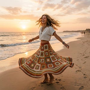 Op de afbeelding: Een vrouw draagt een witte crop top en een lange, kleurrijke gehaakte rok op een strand bij zonsondergang. De rok heeft een patchwork-ontwerp met vierkanten in verschillende kleuren, waaronder oranje, groen en bruin. De rok wappert in de wind.