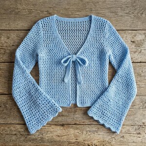 May include: A light blue, open-front crochet cardigan with long, flared sleeves. The cardigan features a V-neckline and a tie closure at the waist. The crochet pattern has a delicate, open weave, and the sleeves have a scalloped edge.
