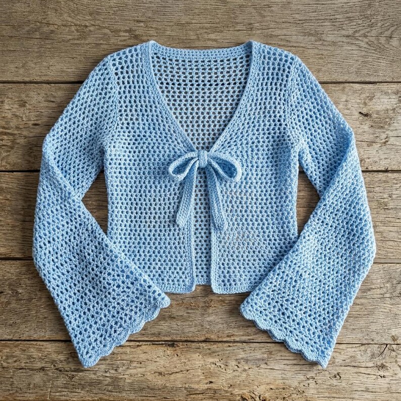 May include: A light blue, open-front crochet cardigan with long, flared sleeves. The cardigan features a V-neckline and a tie closure at the waist. The crochet pattern has a delicate, open weave, and the sleeves have a scalloped edge.