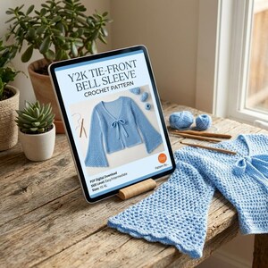 May include: A digital tablet displays a crochet pattern for a Y2K tie-front bell sleeve top. The light blue garment is shown with a bow closure. The tablet rests on a wooden surface, with yarn and crochet hooks nearby. The text on the tablet reads: Y2K TIE-FRONT BELL SLEEVE CROCHET PATTERN.