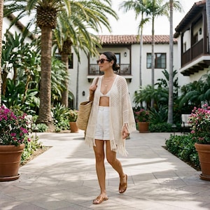 Può includere: Una donna indossa un cardigan all'uncinetto color crema con frange, pantaloncini bianchi e un top bikini bianco. Porta una borsa tote di paglia e sandali e occhiali da sole marroni. L'ambientazione è un vialetto esterno fiancheggiato da palme ed edifici.