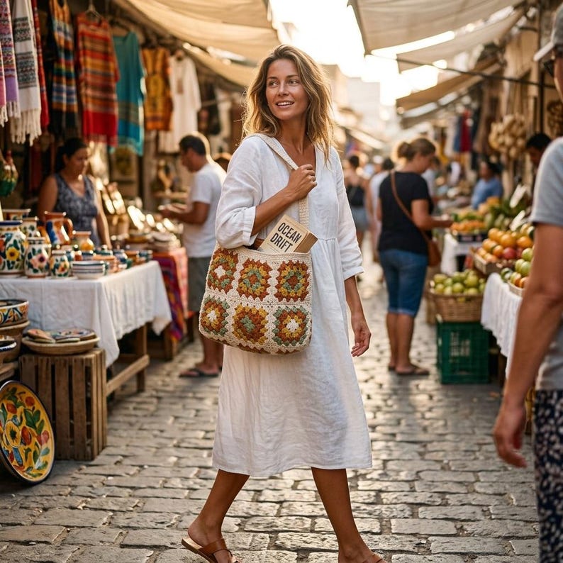 Puede incluir: Una mujer camina por un mercado, llevando un bolso tote de ganchillo. El bolso presenta un dise&ntilde;o de cuadrados de la abuela coloridos en tonos crema, naranja, verde y marr&oacute;n. El bolso tiene una correa color crema y un libro con el texto "OCEAN DRIFT". Lleva un vestido blanco.