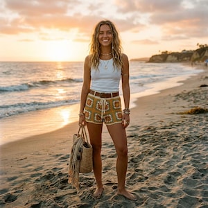 May include: A woman wearing a white tank top, crochet shorts, and a brown belt stands on a beach. The shorts are mustard yellow with a floral pattern. She carries a straw tote bag with a scarf and sunglasses. The background features the ocean and a sunset.
