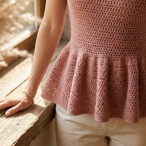 May include: A dusty rose crochet top with a flared peplum. The top is sleeveless and has a textured stitch pattern. The person is wearing a gold bracelet. The top is paired with beige pants.