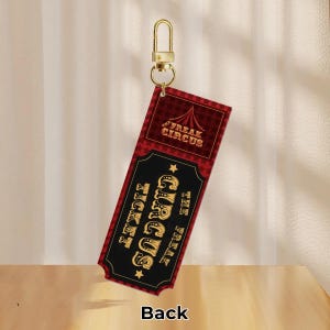 Puede incluir: Un llavero rojo y negro con un dise&ntilde;o de entrada de circo. La entrada dice "Freak Circus" y "The Freak Circus Ticket" en letras doradas. La parte posterior de la entrada es visible y dice "Back". El llavero tiene un cierre dorado.