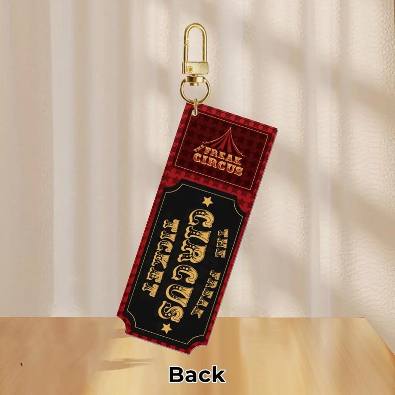Puede incluir: Un llavero rojo y negro con un dise&ntilde;o de entrada de circo. La entrada dice "Freak Circus" y "The Freak Circus Ticket" en letras doradas. La parte posterior de la entrada es visible y dice "Back". El llavero tiene un cierre dorado.