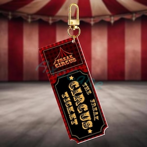 Könnte beinhalten: Ein rechteckiger Schlüsselanhänger mit einem Zirkus-Ticket-Design. Das Ticket hat ein rot-schwarzes Farbschema mit den Worten "FREAK CIRCUS" und "CIRCUS TICKET" in goldener Schrift. Oben ist ein goldfarbener Verschluss befestigt.