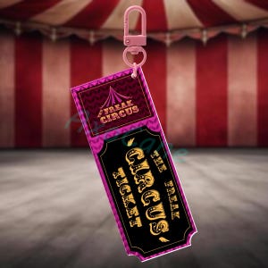 Puede incluir: Un llavero rosa con una etiqueta rectangular. La etiqueta tiene las palabras "The Freak Circus Ticket" en dorado sobre un fondo negro. La parte superior de la etiqueta tiene un dise&ntilde;o rosa y morado con las palabras "Freak Circus".