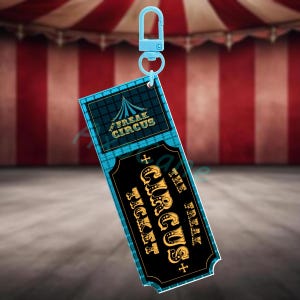 Puede incluir: Un llavero azul con un dise&ntilde;o de boleto "Freak Circus" negro y dorado. El boleto tiene un borde turquesa y un cierre azul. El fondo es una carpa de circo a rayas rojas y blancas.