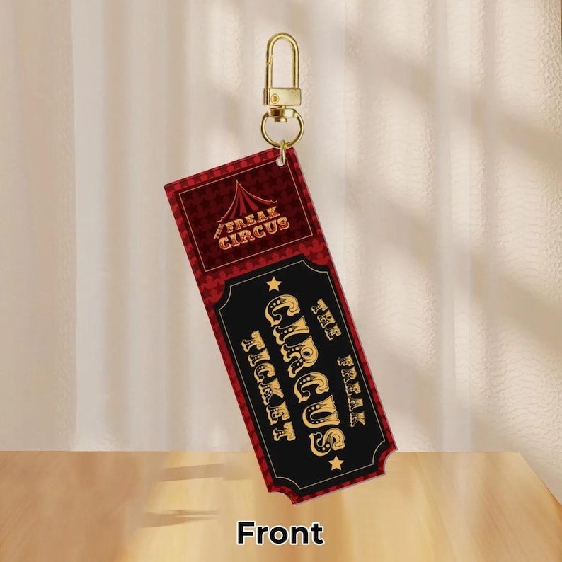 Puede incluir: Un llavero rectangular con un dise&ntilde;o de entrada de circo. La entrada es principalmente negra y roja con letras doradas que dicen "FREAK CIRCUS" y "CIRCUS TICKET". Un cierre dorado est&aacute; adjunto en la parte superior.